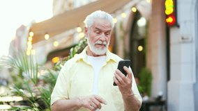 Senior gray haired bearded man talking on a video call using a smartphone while standing on a city street. Happy mature old male in a shirt waving hand, speaks with friend looking at the phone screen