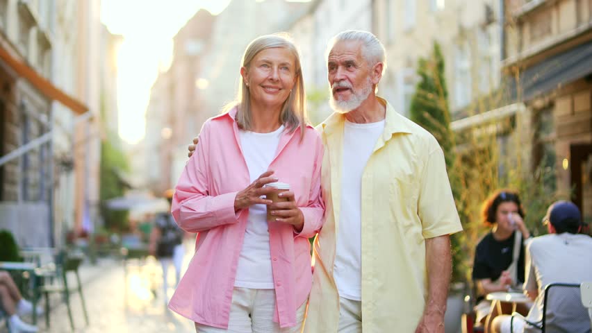 Happy old senior married couple spend leisure time together walking on street in city. Joyful mature gray haired retired wife and husband happily talking, hugging, enjoying a walk downtown outside Royalty-Free Stock Footage #1108515953