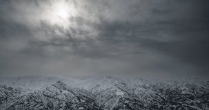 Winter mountains in snow timelapse. Snow-capped mountain ranges on cloudy day. Whole sky is in gray clouds rays of sun are barely visible behind clouds. In some places clouds part and can see blue sky