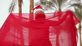 A brunette in red and a santa hat dances an oriental dance against the backdrop of palm trees. Overall plan