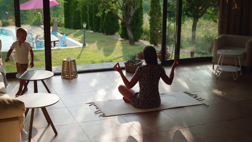 Young woman is trying to meditate on the floor in a luxury house with a view of the pool in the yard, sitting in lotus pose, and her 3-year-old son running around her.