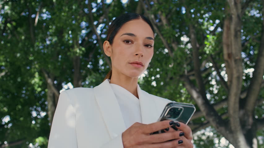 Portrait of confident woman ceo using mobile phone standing under tree. Closeup attractive serious businesswoman in stylish white suit texting smartphone outdoor. Chic lady posing camera in green park