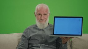 Green Screen.Portrait of an Old Man with a Laptop with Blue Screen Shows Thumb Up, Sitting on a Sofa. Smart Homes and the Internet of Things for Seniors.