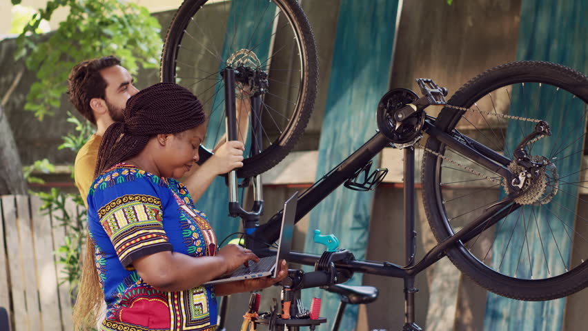 Vibrant african american lady browsing on internet for bicycle gear maintenance. Black woman checking for repair instructions on laptop while caucasian man dismantles broken bike wheel. | Shutterstock HD Video #1108525243