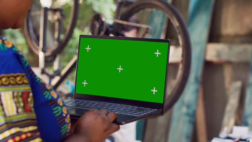 Detailed view of african american lady grasps minicomputer with greenscreen display as man in background removes broken wheel. Black woman holds laptop with blank mockup screen for bike maintenance. | Shutterstock HD Video #1108525257