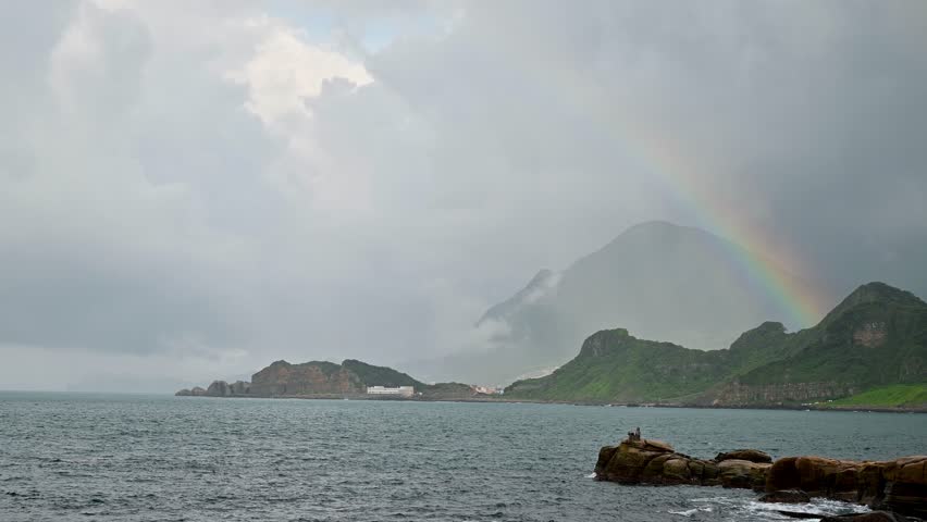 There is a rainbow in the sky by the sea. Rocky coast under blue sky and white clouds. White waves and light blue sea.