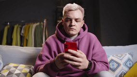 Fair-haired guy with phone on sofa in living room-studio. Neon lights in the background. Expression of emotions, dissatisfaction, disappointment, gets up and leaves, throwing phone on sofa. Losing