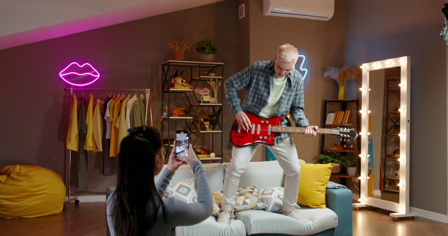 Video for social media guy. Shooting on smartphone, shorts, reels, stories. He plays electric guitar, having entered courage and jumping to beat on couch, neon lights, home studio. Virtual lifestyle | Shutterstock HD Video #1108530149
