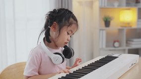Asian cute girl smile and wearing white headphone playing learning online piano music in the living room at home. The idea of activities for the child at home during quarantine. Music learning study.