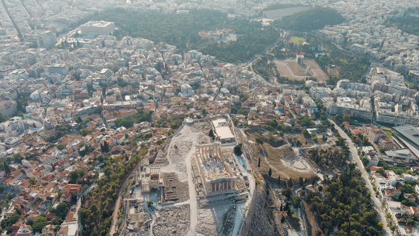Athens, Greece. The Acropolis of Athens rises on a rocky plateau above the dense urban grid of the modern city, captured in the warm, diffused light of early morning during summer. The Parthenon, a Doric temple of white marble with its peristyle of fluted columns, stands at the center of the citadel, surrounded by ancient walls, ruins, and the remains of other classical structures. The camera maintains a steady aerial perspective, slowly pulling back to reveal the full extent of the fortified hill, the Theatre of Dionysus at its southern base, and the sprawling cityscape extending toward hazy mountains on the horizon. The scene is bathed in soft golden light with long shadows, emphasizing the contrast between the pale stone of the archaeological site and the surrounding greenery and urban development. The atmosphere is serene and monumental, conveying the enduring presence of one of the most significant ancient sites in the world. The Parthenon was constructed almost entirely without straight lines, its architects employed subtle optical refinements, including a curved stylobate and inward-leaning columns with entasis, to create an appearance of perfect geometric harmony to the human eye.