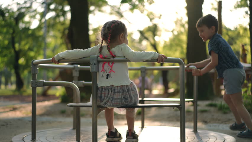 Joyful boy running spinning merry-go-round with little girl inside. Cheerful carefree Caucasian happy friends enjoying leisure in spring summer park at sunset outdoors