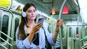 Female chatting online in mobile phone messenger watch video in metro. Young woman passenger standing with headphones and smartphone while moving in modern tram, enjoying trip at the public transport.