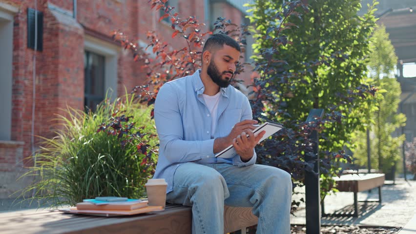 Focused indian student looking for inspiration outdoors and sitting on street bench with notebook in hands. Thoughtful man with cup of coffee-to-go dressed in blue jeans and shirt writing essay.