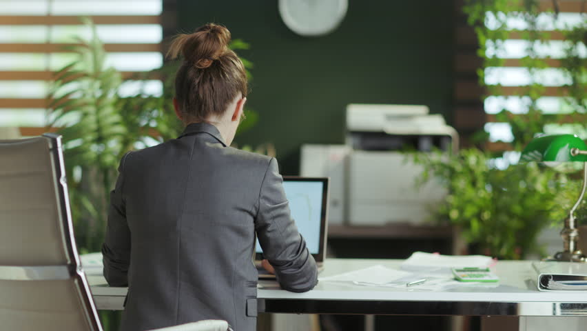 Sustainable workplace. Seen from behind modern business woman in a grey business suit in modern green office stretching neck. Royalty-Free Stock Footage #1108544231