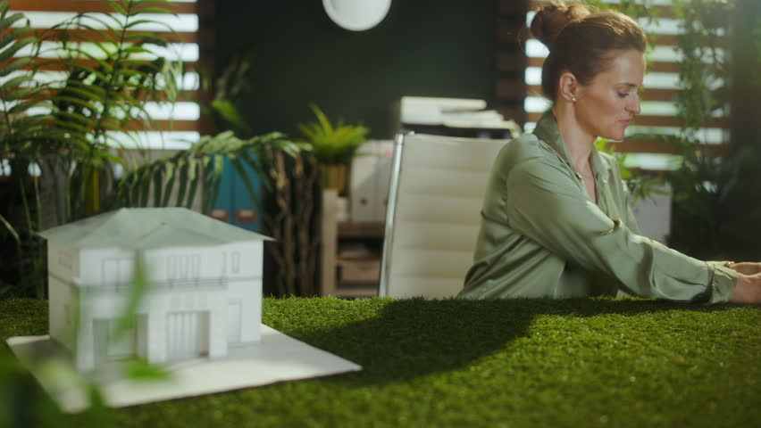 Eco trends. happy middle aged business woman in green blouse in modern green office with paper house and windmill.