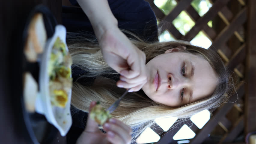 A girl in a blue dress eats snails with cheese with a fork with her hands at a wooden summer table and wipes her hands with a napkin. vertical video | Shutterstock HD Video #1108547461