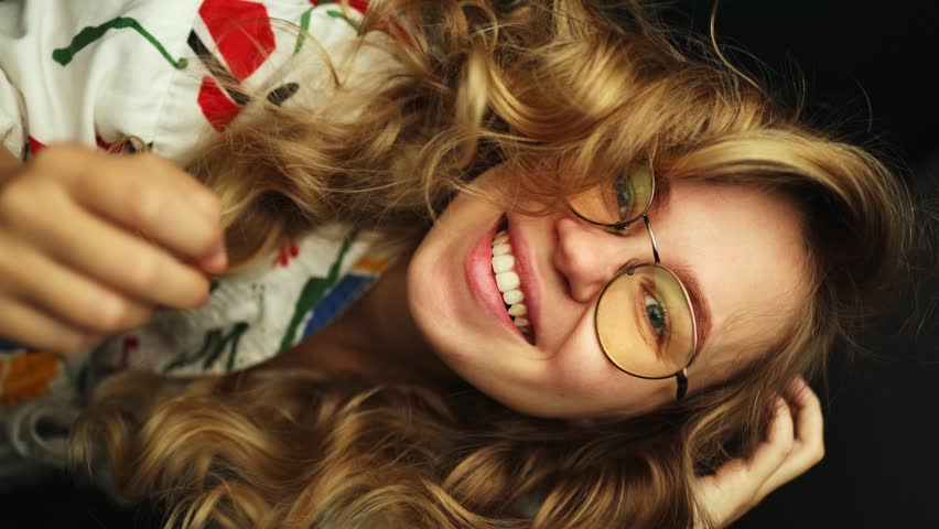 A beautiful girl in a bright colored robe poses for the camera and adjusts her glasses and her curly hair. girl emotionally plays on the camera at home on a black background. vertical video | Shutterstock HD Video #1108547473