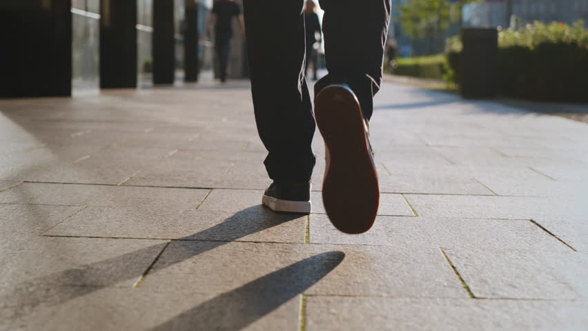 Back view following male feet steps walking busy street on sunny evening. Man wearing casual dark trousers leather sneakers goes on urban sidewalk in city. Low angle people legs backlit warm sunlight Royalty-Free Stock Footage #1108547505