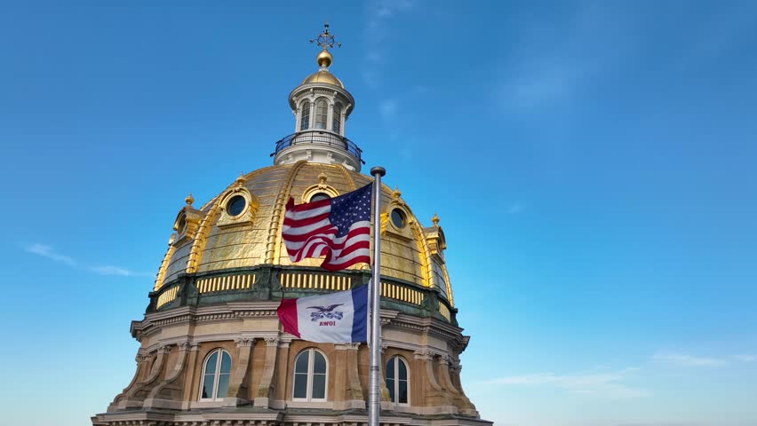 America and Iowa flags waving in front of capitol dome in Des Moines, IA. Cinematic aerial establishing shot.