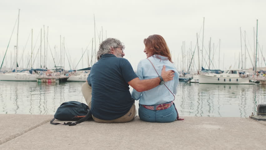 Elderly couple in love talking to each other while sitting in the marina at the pier, back view