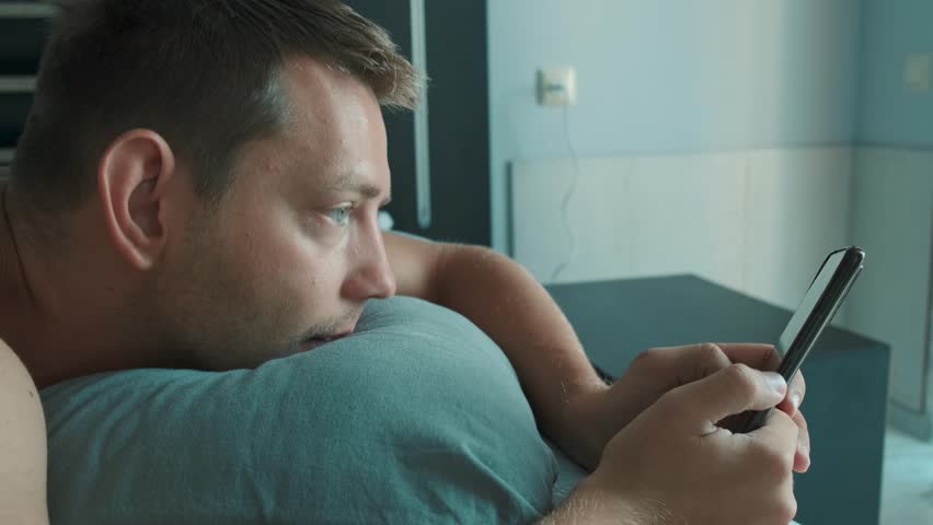 Handsome man using mobile phone in bed at the morning surfing internet, checking social media and reading news. Gadget addiction. Technology concept
