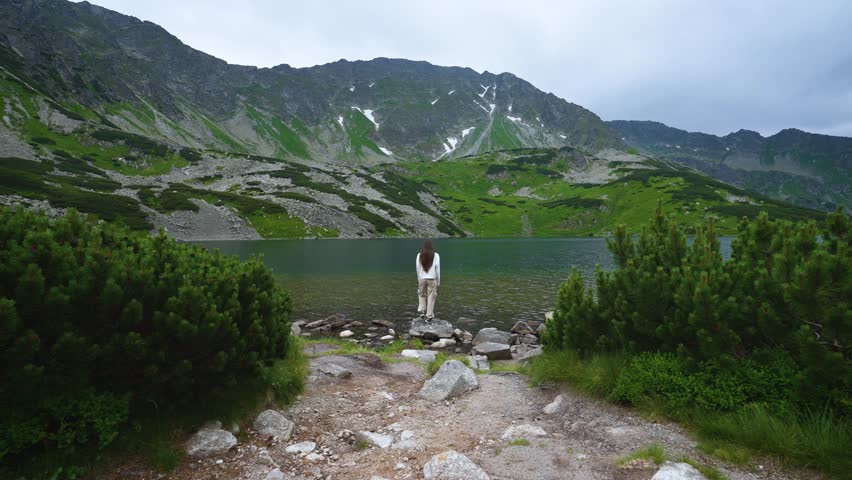 Beautiful mountain landscape of a young long-haired woman or girl standing on a stone on the shore of a mountain lake and enjoying the view spreading her arms to the sides and raising them