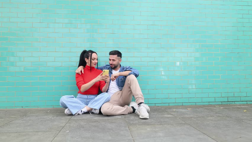 young adult couple sitting on the floor while taking a selfie in front of colorful background