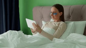 Beautiful young woman with patches on her eyes is lying on the bed in the bedroom at home and using a tablet