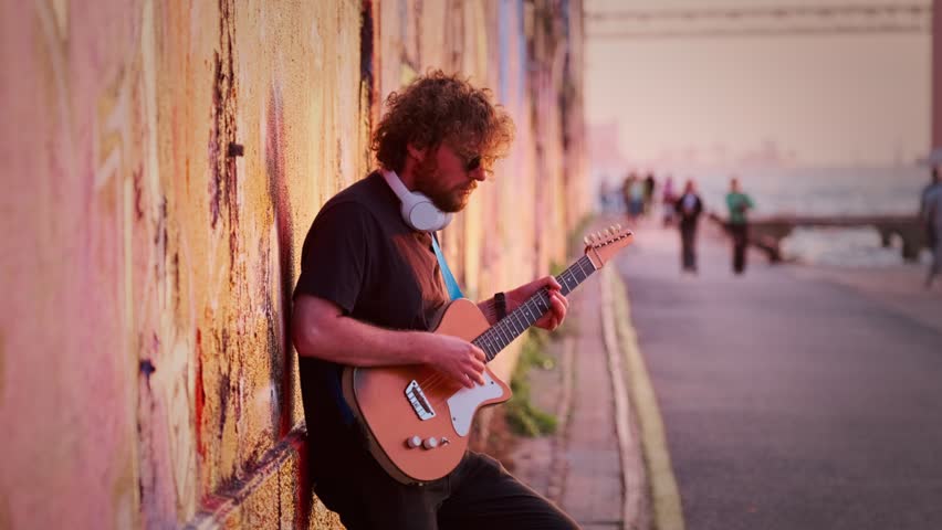 Hipster street musician in black playing electric guitar in the street on sunset leaning on a painted wall