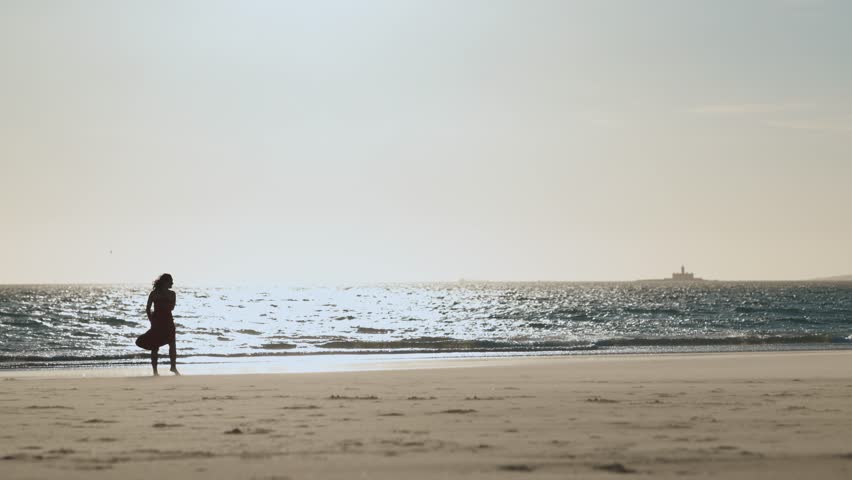 Graceful young woman in red dress dancing alone on the beach at early sunset
