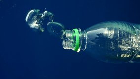 Vertical video, Air bubbles come out of discarded plastic bottle and it slowly sinks into blue abyss in rays of sunlight. Transparent plastic bottle descend to depth blue of sea, Close up, Slow motion