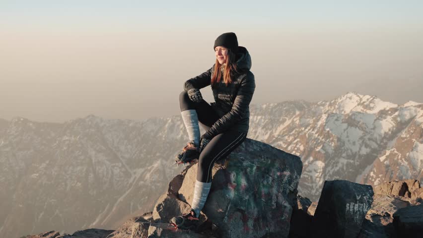 Young caucasian woman sitting on a rock on top of Mount Toubkal, with mountain range in the background, Morocco.