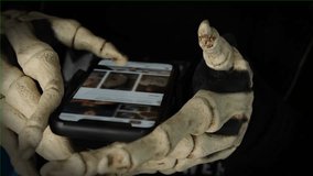 Scary hands in skeleton costume hold smartphone with chromakey scrolls through the social media feed, dark vignette, close up. Virtual dance parties, ghost stories, supernatural sightings.