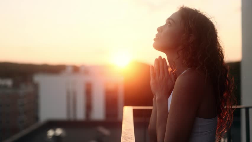 Portrait of hopeful young curly woman clasping hands in prayer asking for blessing and help while the rays of rising sun fall on her face at balcony outdoors Religion and faith concept Royalty-Free Stock Footage #1108599495