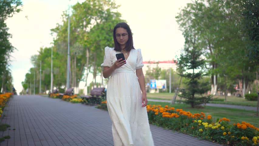 A young woman walks along a flower alley with a phone in her hands. A girl in a white dress looks at her phone and walks among the flowers.