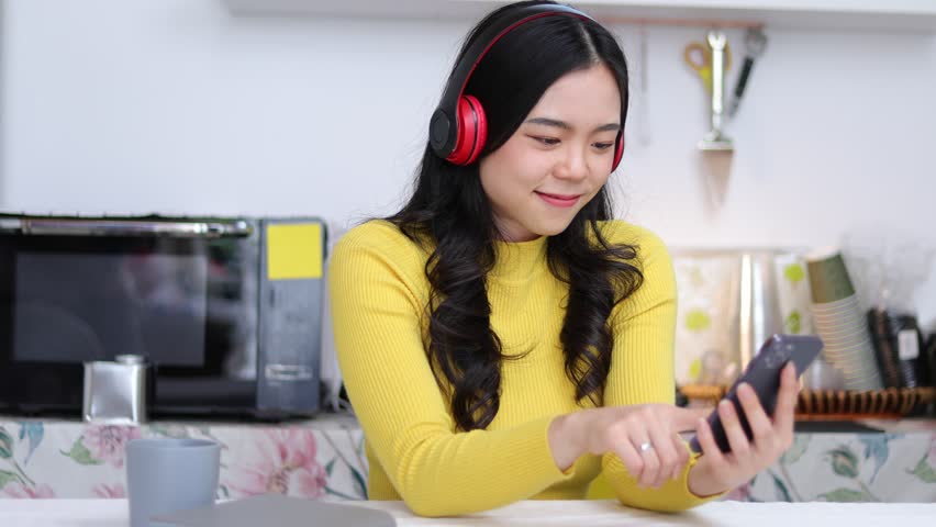 Asian woman happy at home wearing headphones listening to music on mobile.
