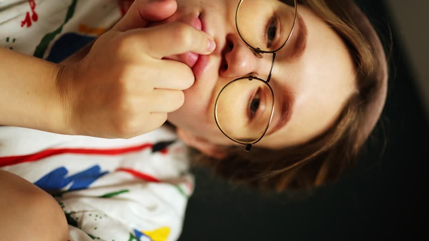 Pensive girl in glasses and a dressing gown holds her hand on her chin and looks at the camera and smiles. vertical video. adjusts glasses and flirts | Shutterstock HD Video #1108600889