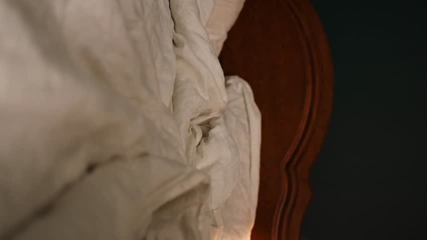 A girl in a bright bathrobe and glasses sits on the bed and covers herself with a white blanket and takes off her glasses. vertical video. | Shutterstock HD Video #1108600901