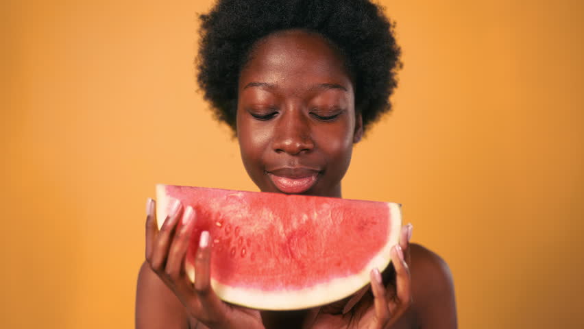African American young woman with afro hair takes a tasty bite of juicy watermelon isolated on orange background. Skin care. Healthy lifestyle.