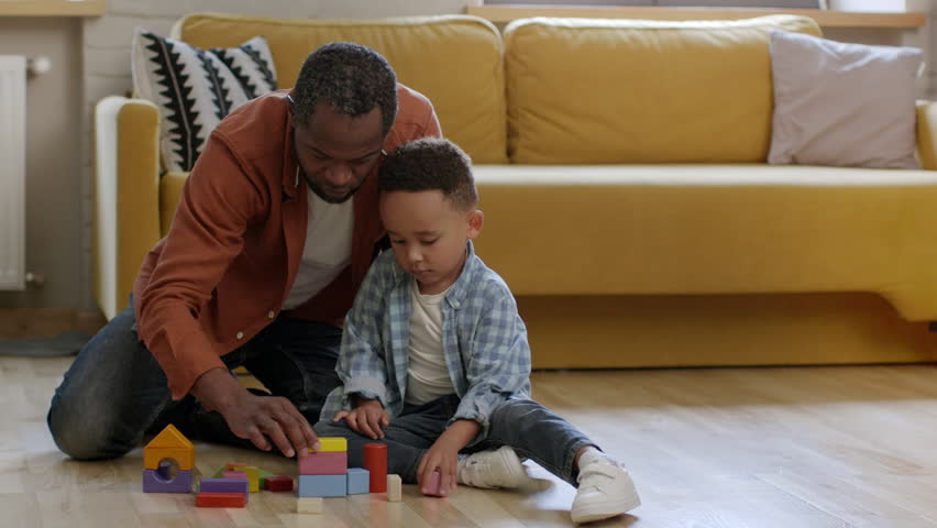 Paternity leave and kids development. Loving african american daddy playing colorful blocks with his cute little son, building castle together on floor at home, tracking shot, slow motion Royalty-Free Stock Footage #1108616139