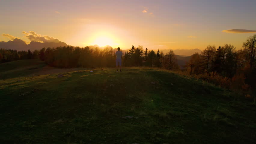AERIAL: Young man jumps with raised arms when he reaches top of a hill at sunset. He celebrates his hiking achievement at picturesque peak, surrounded by wonderful views of mountains on an autumn day.