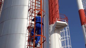 Heating Plant Worker Climbing Ladders of Industrial Storage Tank. Maintenance Technician Climbing up a Metal Staircase at District Heating Plant. Industrial Chimneys and Storage Tanks Against Blue Sky - Powered by Shutterstock - Get 15% off with code: PIKWIZARD15