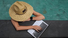Female freelancer in straw hat working with laptop in the swimming pool at sunny day. High quality 4k footage