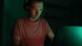 An adult male gamer in headphones plays on a laptop in a dark room.