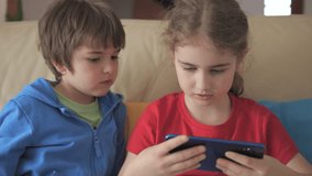 Children Playing Games In Phone at Home on Couch. Kids Playing Video Game on Mobile Phone. Boy and Girl Plays Video Game Smartphone on Sofa Friends Using Phone for Gaming Online Education Social Media