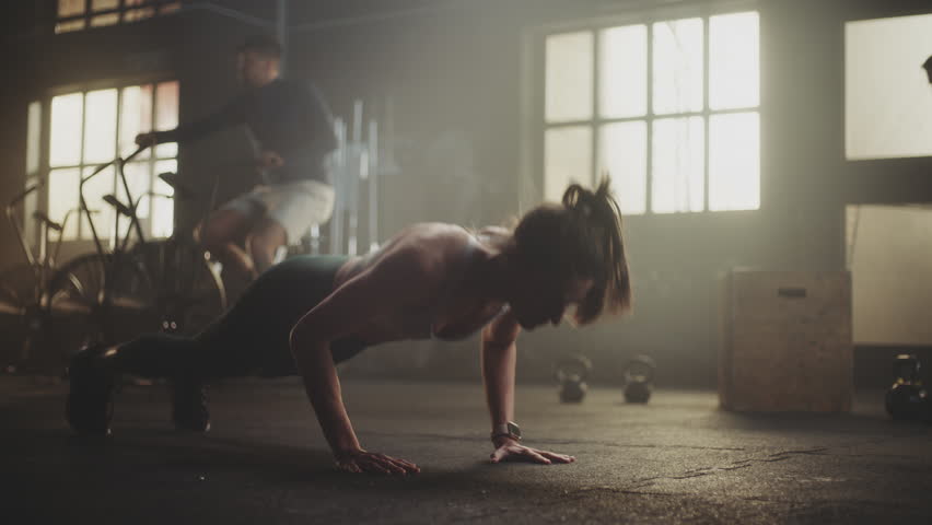 Attractive young woman wearing sports bra doing pushups at a gym while two other men exercising in the background. Slow motion handheld medium shot Royalty-Free Stock Footage #1108631935