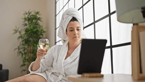 Young beautiful hispanic woman wearing bathrobe drinking champagne using touchpad at bedroom