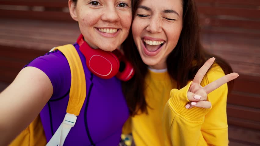 Portrait of a brunette girl in a yellow sweater and a girl with a short haircut in a purple top and red headphones who take a selfie on a brown bench in a skatepark in summer