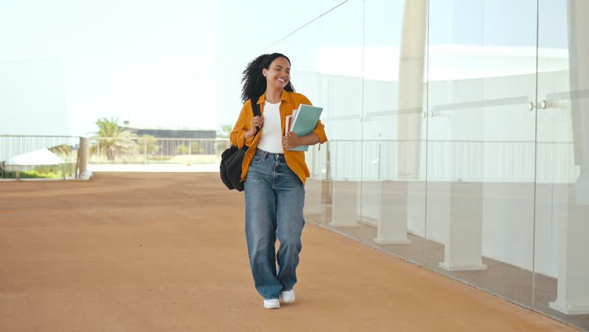 Full-length photo of cheerful amazing curly haired mixed race female student, with a backpack, hold books, notebooks in her hand, walking near the university, looks away and smile, finished school day Royalty-Free Stock Footage #1108645015