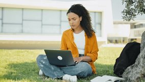 Full-length video of a upset disappointment brazilian or hispanic curly haired girl, freelancer, sitting on the grass near the tree, with a closed laptop, feels anxious, frustrated, sadly looks away